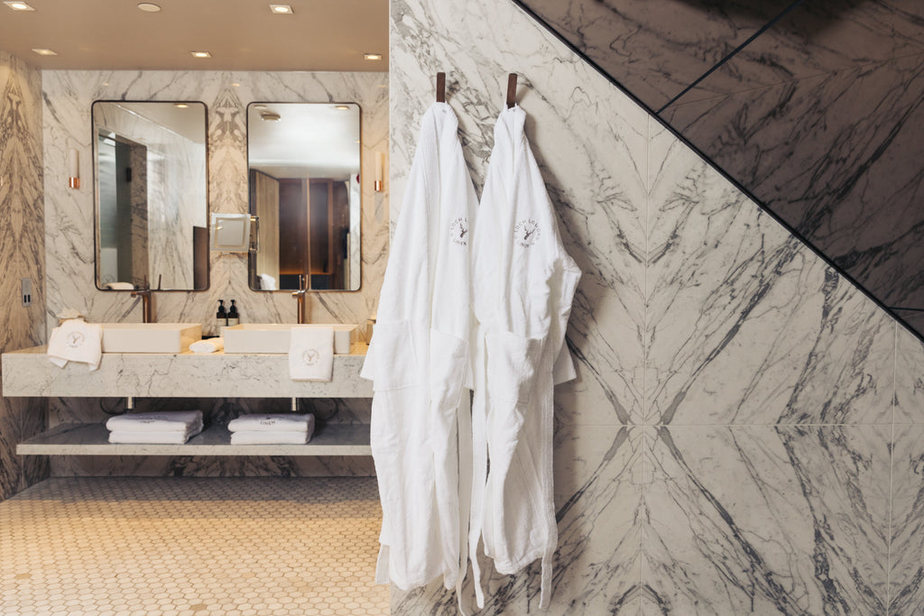 Two luxury 100% cotton bathrobes are hung in an elegant marble walled bathroom. Loch Lomond Linen's white dressing gowns are unisex, suitable both for women and men.