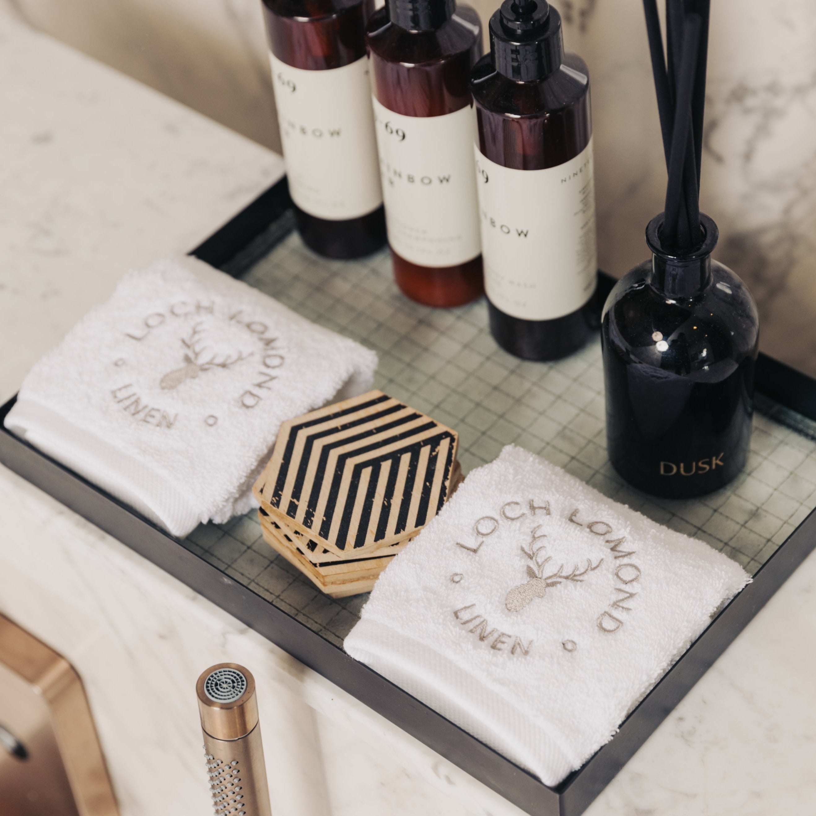 Two luxurious white 100% cotton face towels from Loch Lomond Linen is photographed in an elegant hotel bathroom with marble walls. The face towels are neatly folded into squares and placed next to some skincare products.