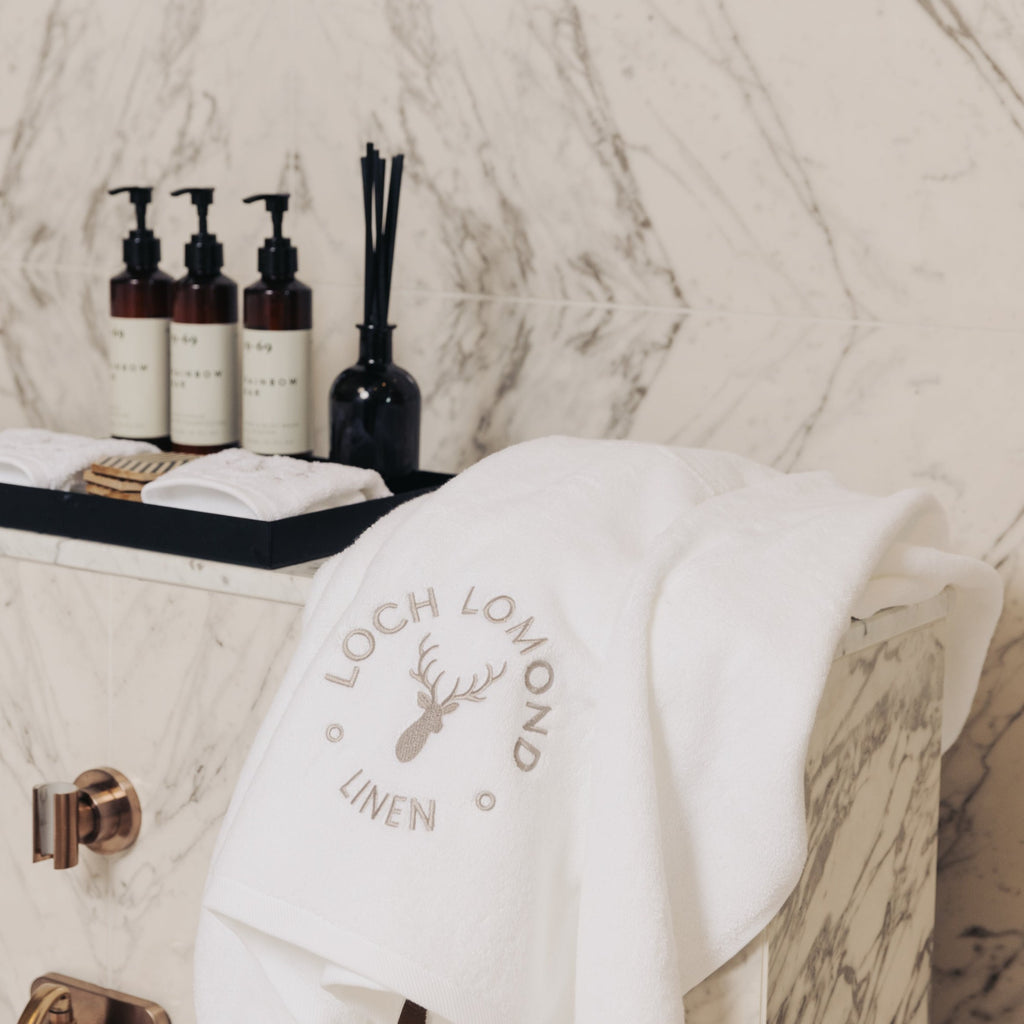 A luxurious white 100% cotton bath towel from Loch Lomond Linen is photographed in an elegant hotel bathroom with marble walls.