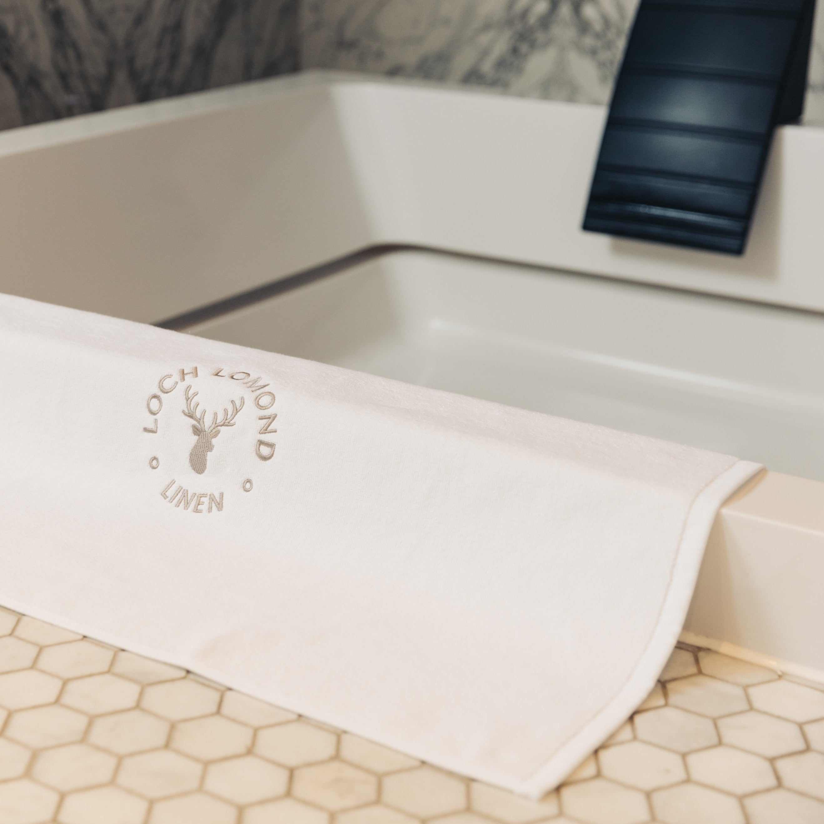 Loch Lomond Linen's luxurious white 100% cotton bath mat is photographed at the edge of an elegant bath tub.