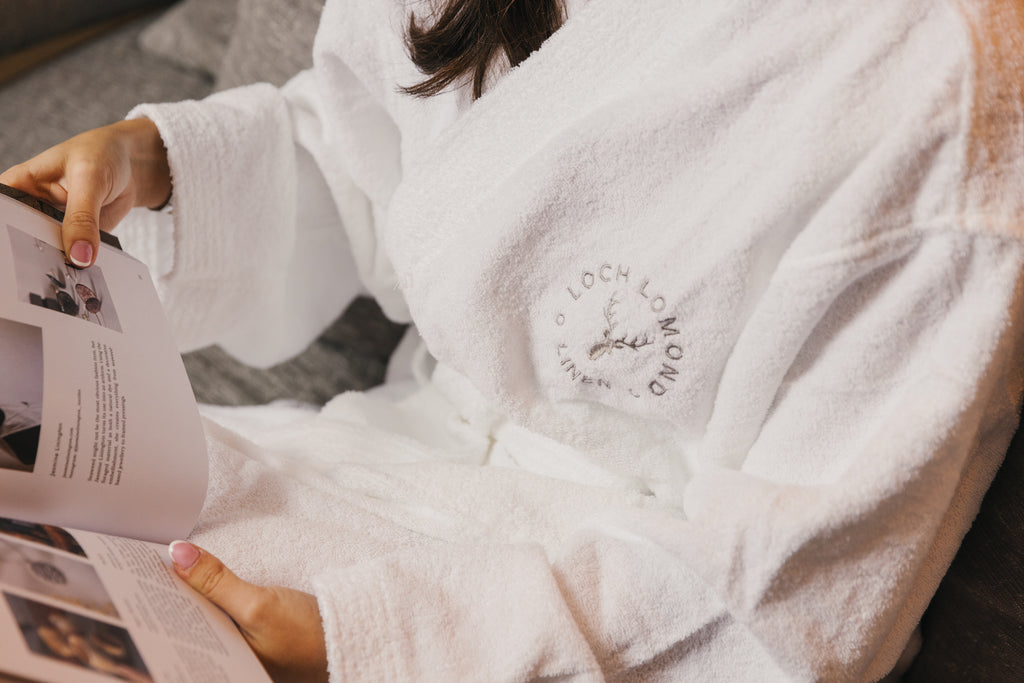 A brown haired woman is wearing Loch Lomond Linen's luxury white terry towelling dressing gown while reading a magazine. The brand's logo is embroidered on the front of the bathrobe. The robe is suitable for both women and men.