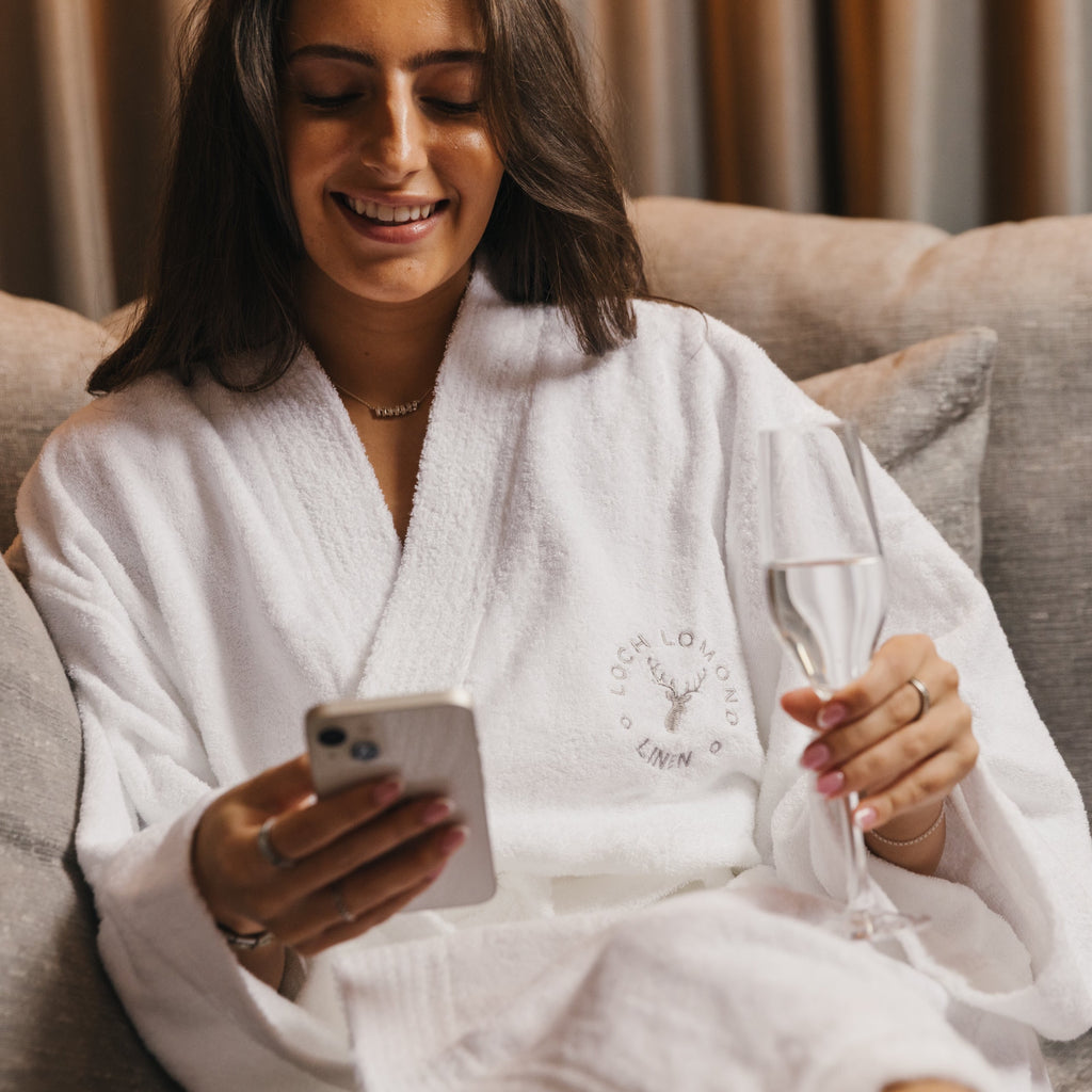 A tanned, brown haired woman is wearing a white pure 100% cotton terry towelling dressing gown from Loch Lomond Linen in an elegant hotel room and drinking chanmpagne while looking at her phone and smiling.