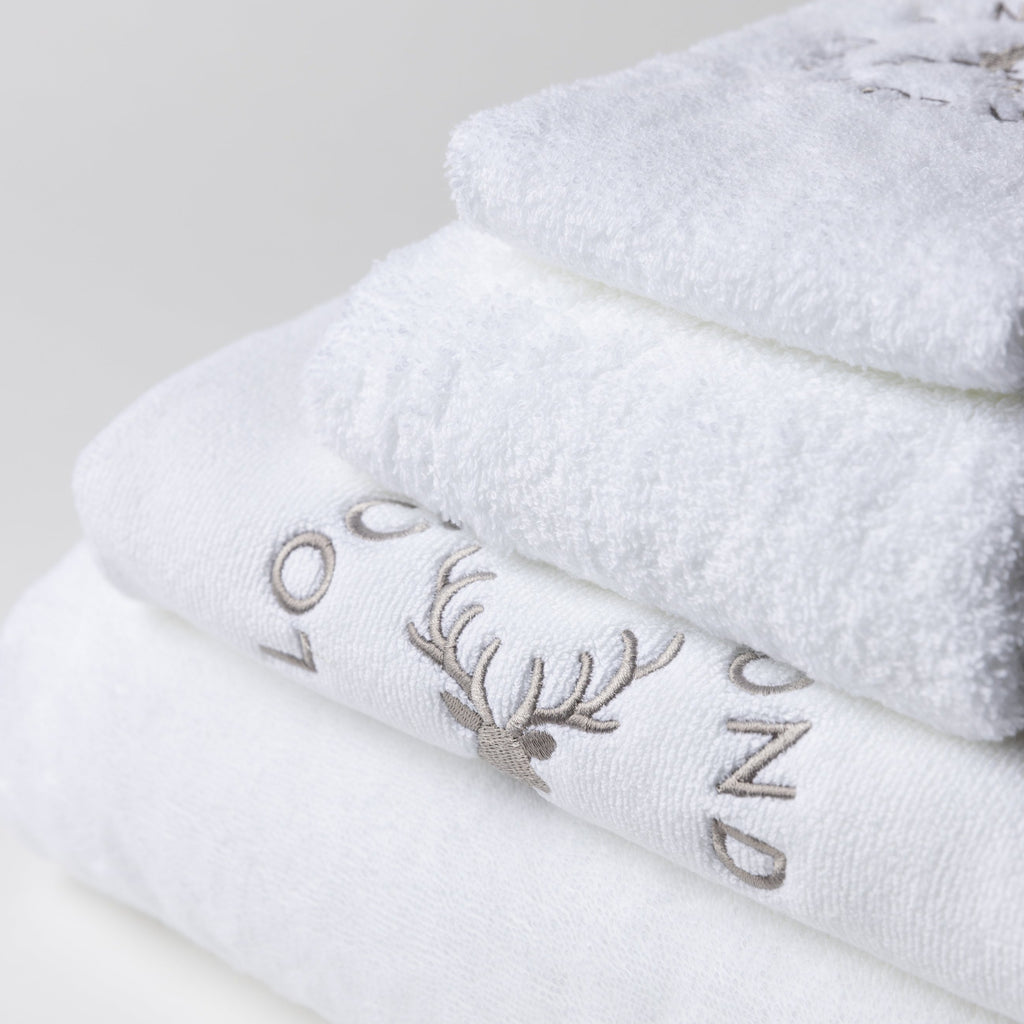 The three white cotton towels and bath mat set is shown from close up. The products are bright white coloured and look extremely soft. The grey Loch Lomond Linen logo is embroidered onto the products.