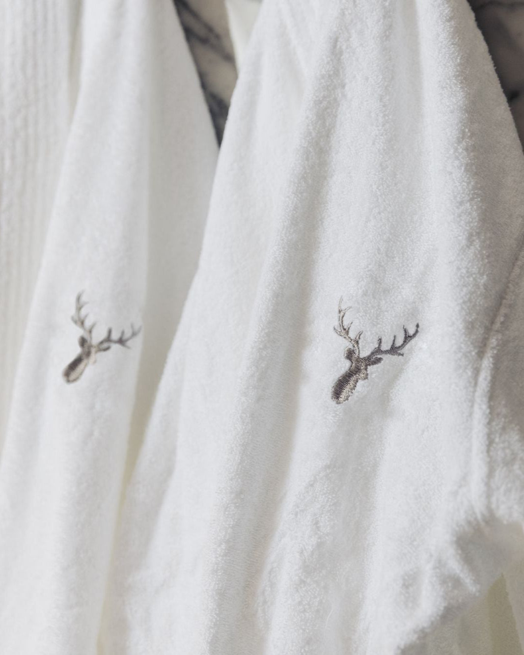 Two 100% cotton hotel-style quality bathrobes are hanged in a bathroom. Loch Lomond Linen's logo is embroidered on the front. The robes are unisex, suitable for men and women alike.