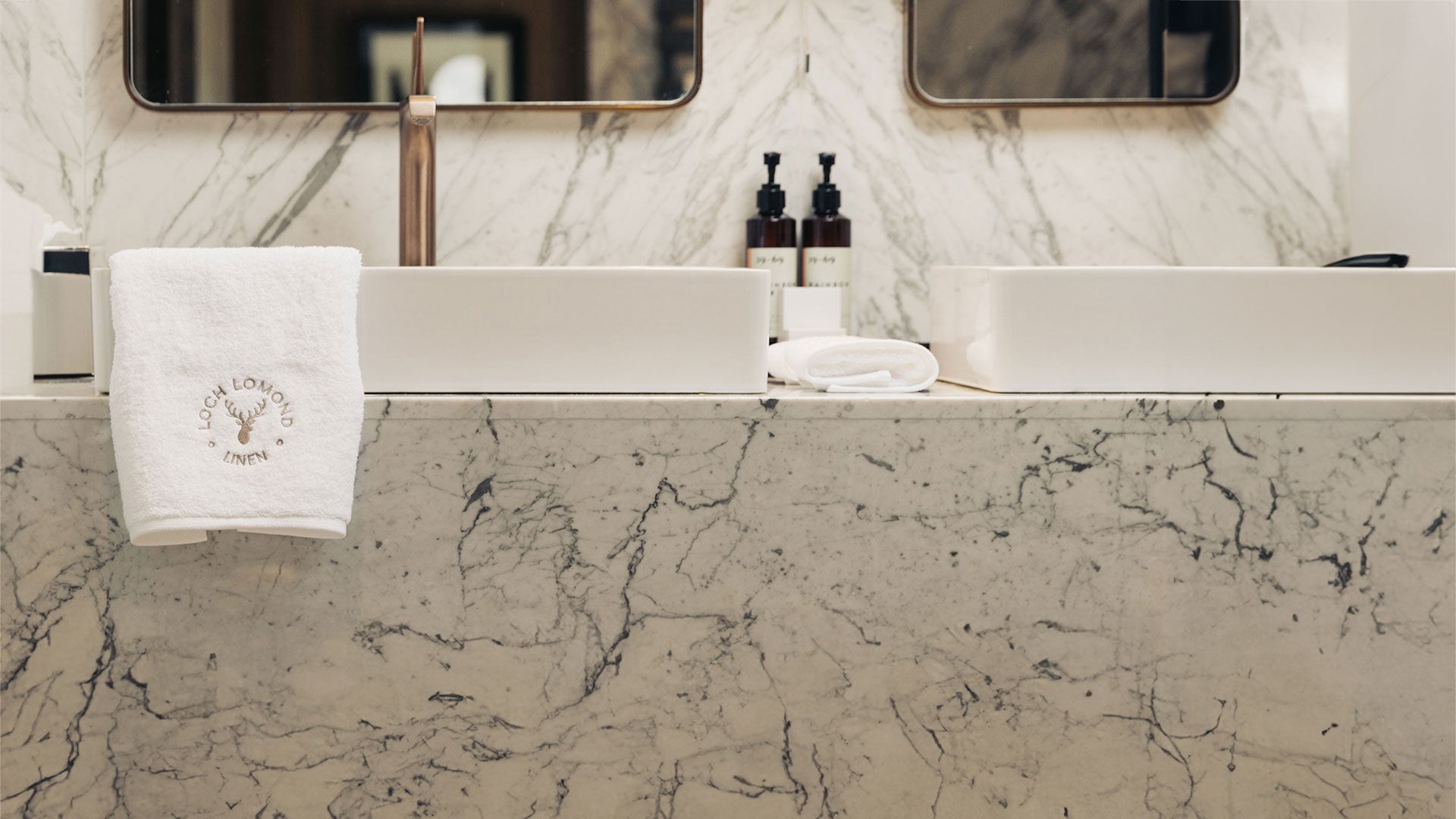 Luxury white 100% cotton towels from Loch Lomond Linen shown in an elegant marble walled bathroom around the sinks.