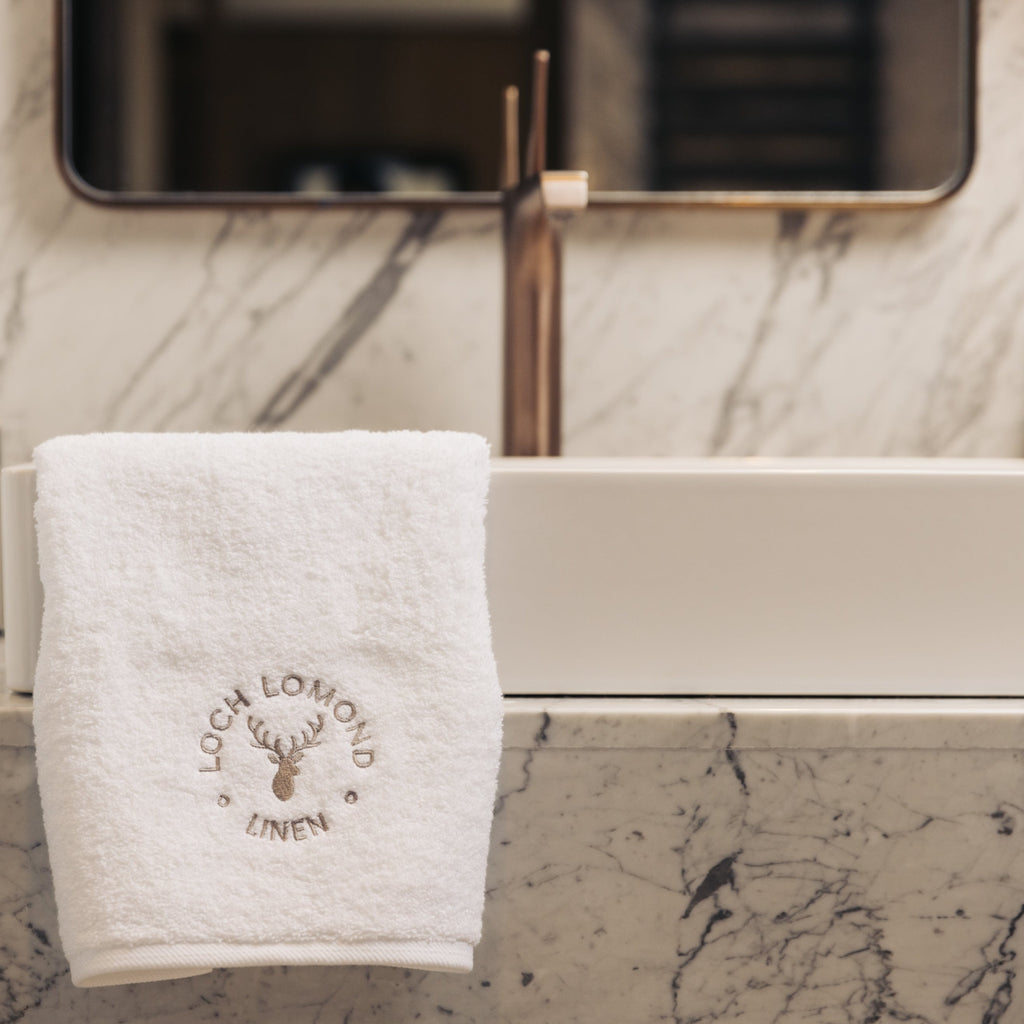 A luxurious white 100% cotton hand towel from Loch Lomond Linen is photographed in an elegant hotel bathroom with marble walls. The hand towel is elegantly layed onto the sink. The brand's sophisticated logo is shonw on the product.