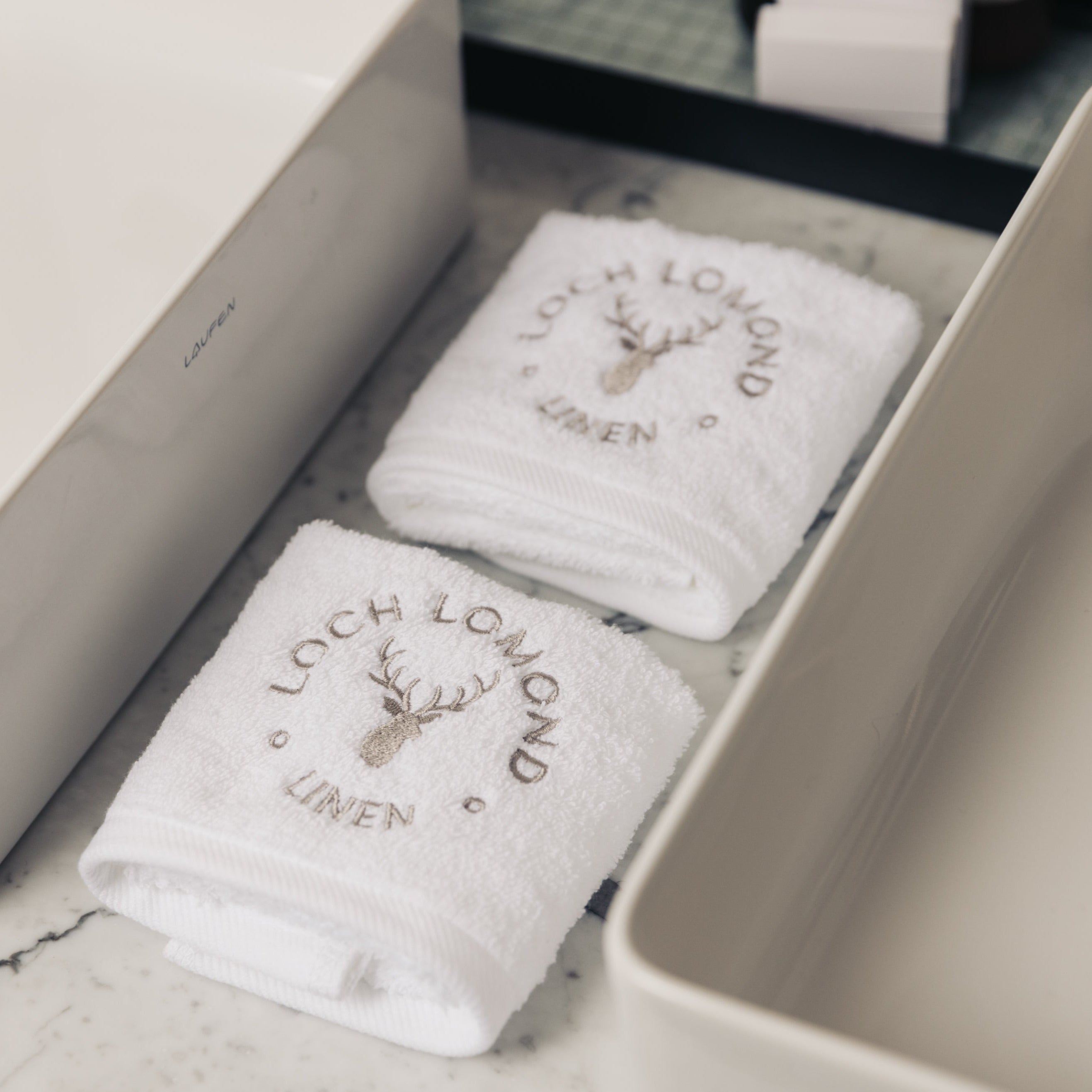 Two luxury face towels with the Loch Lomond Linen logo embroidered on top of them are photographed neatly folded into squares and placed in between two sinks.