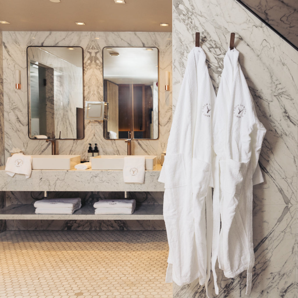 An elegant and sophisticated marble walled bathroom is shown with Loch Lomond Linen's luxury white terry towelling dressing gowns for men and women are hanged on the wall while towels from the same brand are placed under and next to the sinks in the background.