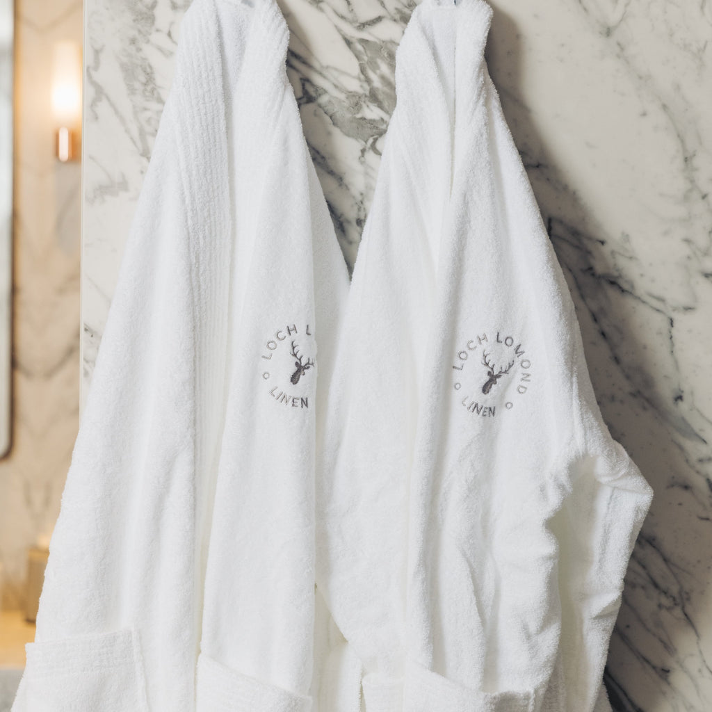 Two white luxurious Loch Lomond Linen bath robes are hanged in a marble bathroom. They are suitable for men and women.