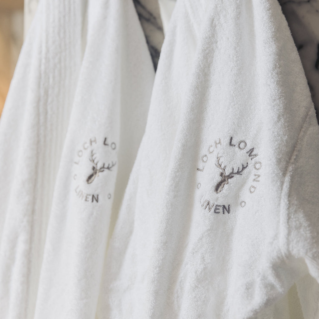 Two 100% cotton hotel-style quality bathrobes are hanged in a bathroom. Loch Lomond Linen's logo is embroidered on the front. The robes are unisex, suitable for men and women alike.