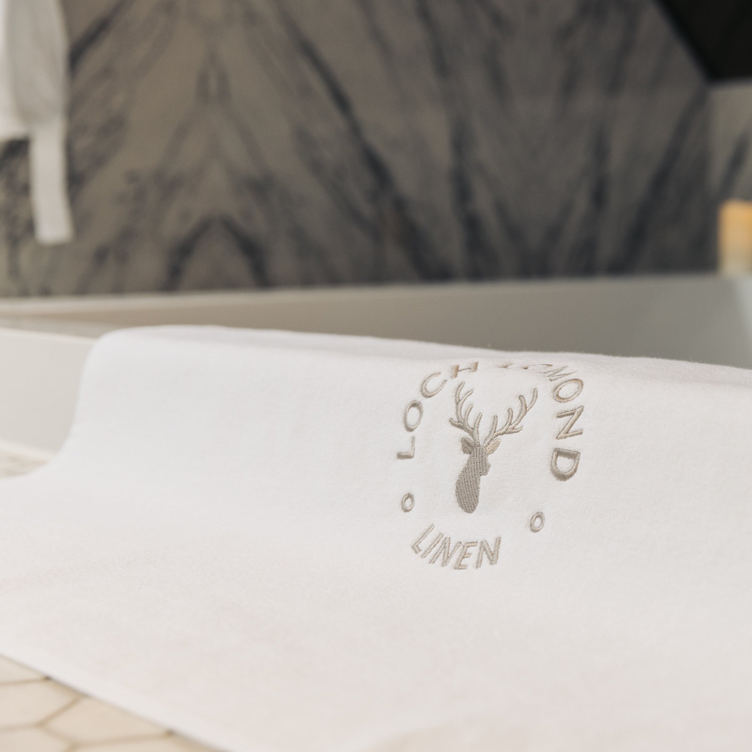 A luxury white cotton bath mat is shown with Loch Lomond Linen's grey logo embroidered in the middle.