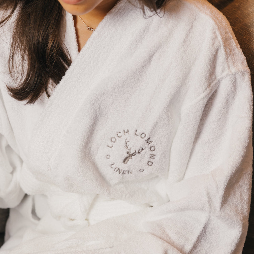 Brown haired woman is wearing Loch Lomond Linen's adult terry towelling bathrobe in white with the grey logo embroidered on the front.