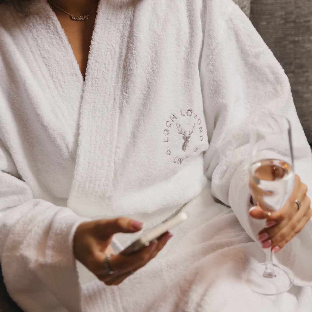 A woman is wearing Loch Lomond Linen's robe while holding a glass of champagne in her one hand and a phone in her other hand.