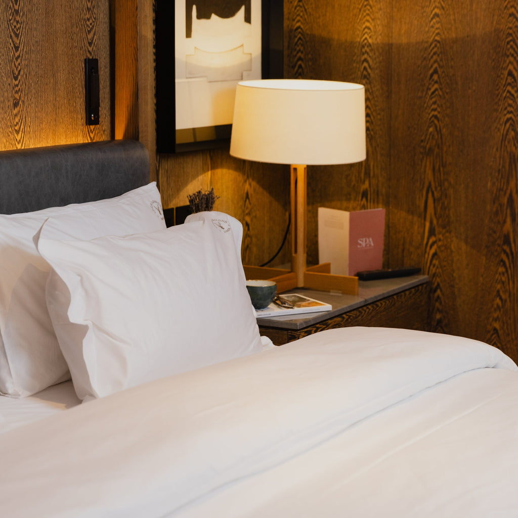 Loch Lomond Linen's white cotton full bedding set is shown on an elegant hotel room’s bed. The brand's logo is embroidered onto the pillowcases.