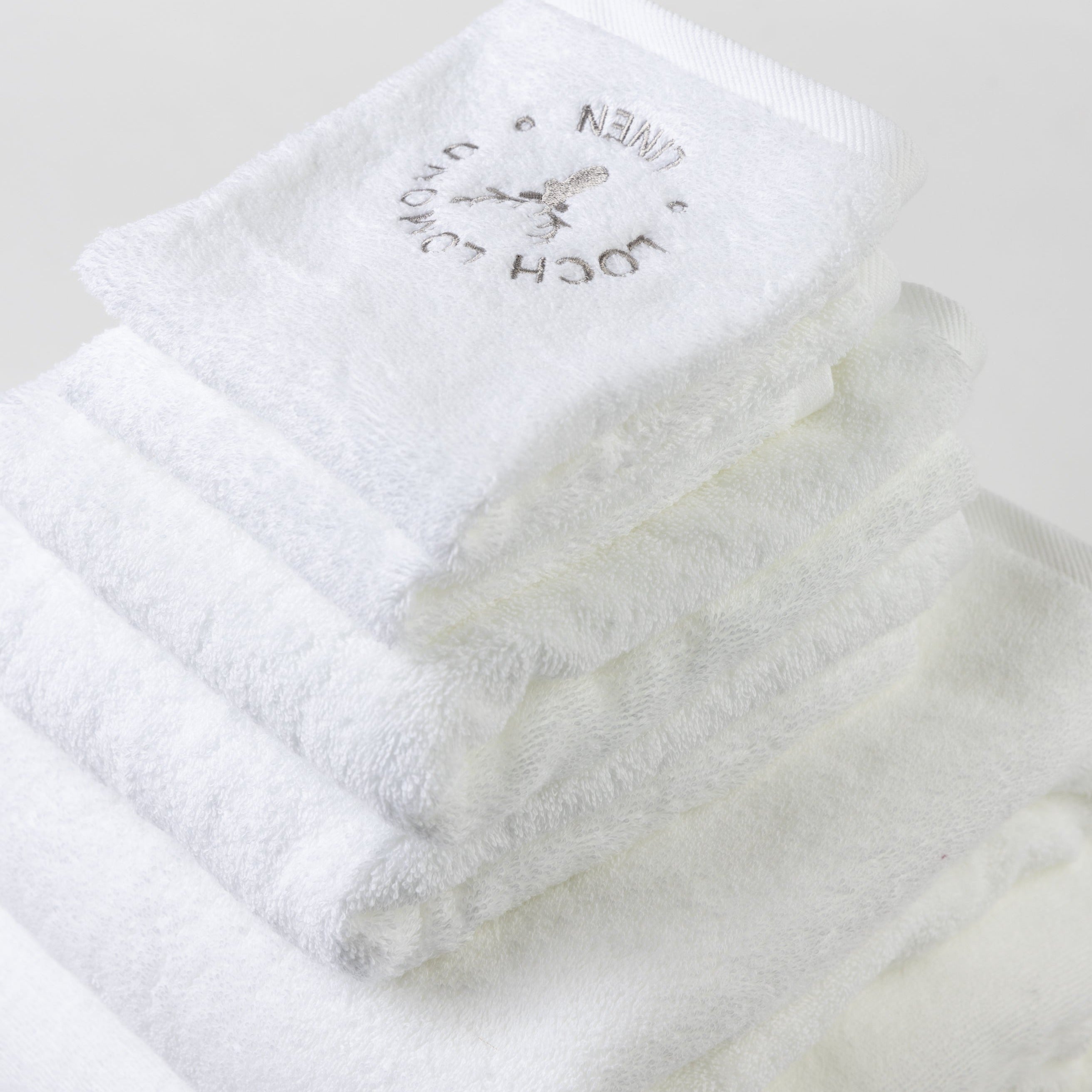 One bath sheet, two bath towels, two hand towels and two face towels are folded into a tower and photographed from the top. The grey Loch Lomond Linen logo is elegantly embroidered on the products.