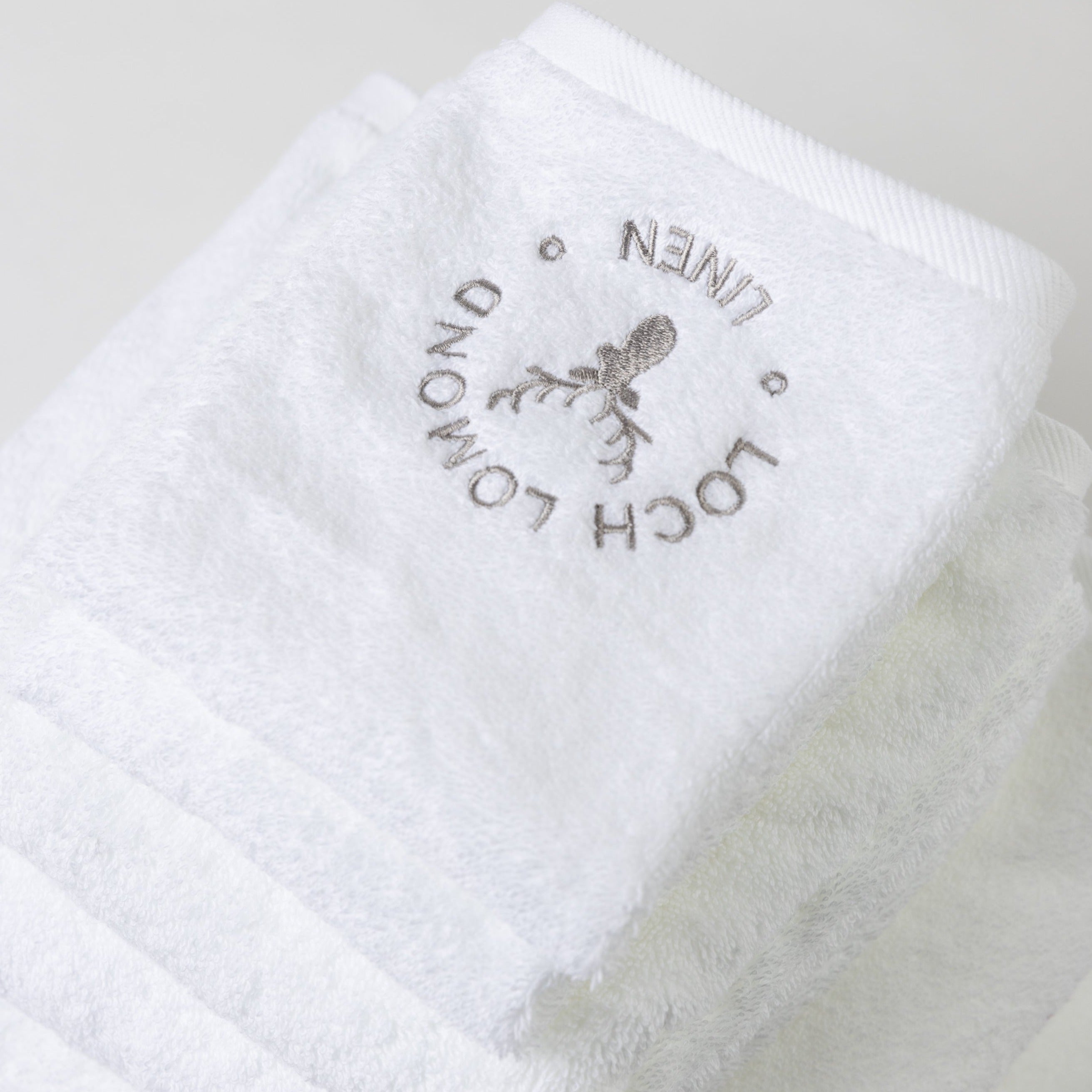 A set of seven luxury 100% cotton white towels are shown withe elegant grey Loch Lomond Linen logo embroidered on the top product.