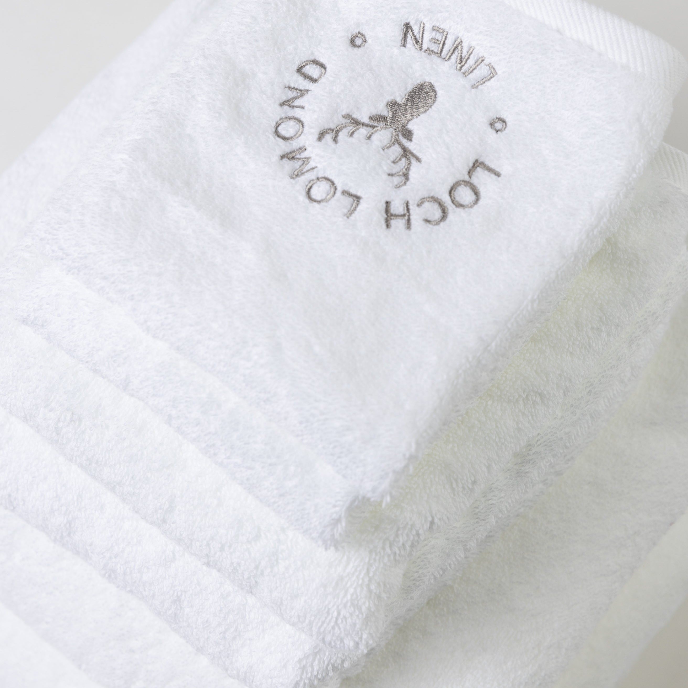 A set of six luxury 100% cotton white towels are shown withe elegant grey Loch Lomond Linen logo embroidered on the top product.