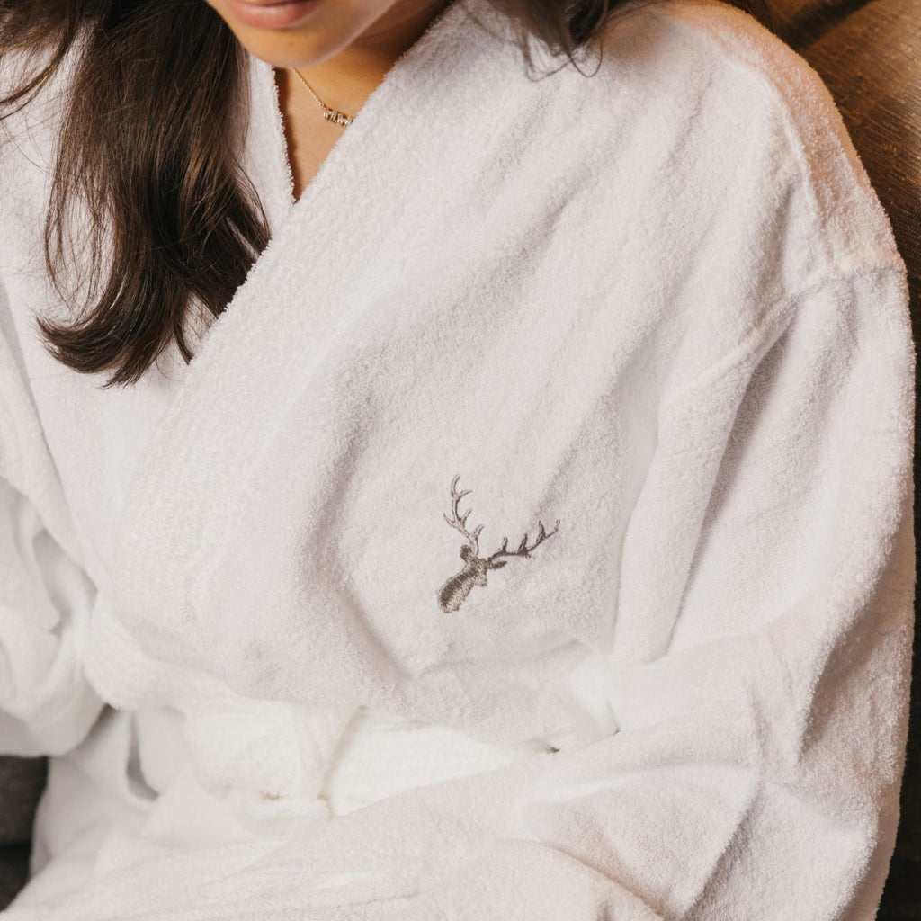 Brown haired woman is wearing Loch Lomond Linen's adult terry towelling bathrobe in white with the grey logo embroidered on the front.