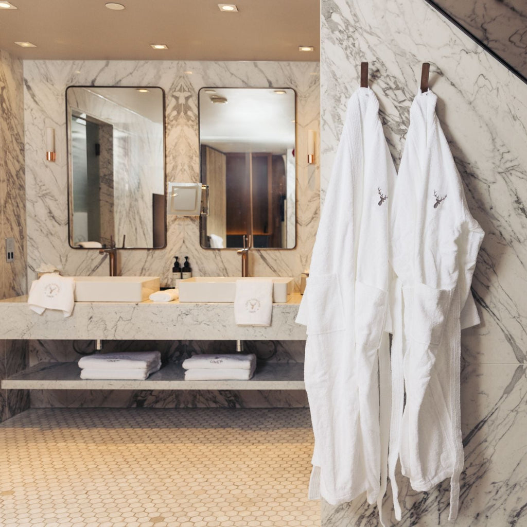 An elegant and sophisticated marble walled bathroom is shown with Loch Lomond Linen's luxury white terry towelling dressing gowns for men and women are hanged on the wall while towels from the same brand are placed under and next to the sinks in the background.