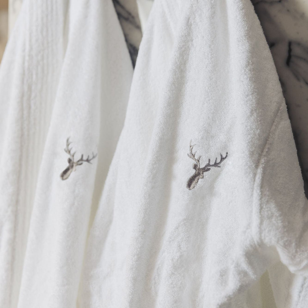 Two 100% cotton hotel-style quality bathrobes are hanged in a bathroom. Loch Lomond Linen's logo is embroidered on the front. The robes are unisex, suitable for men and women alike.