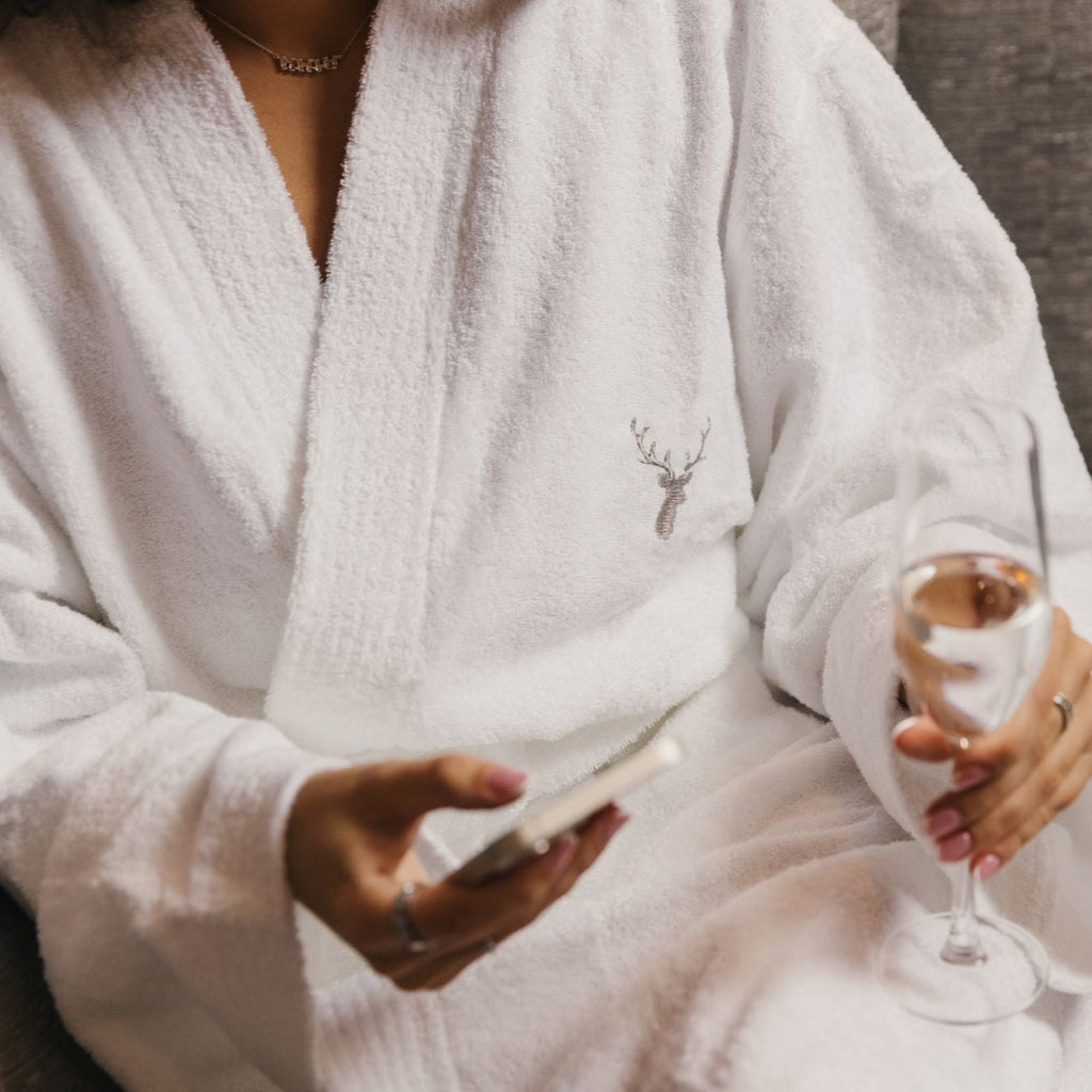 A woman is wearing Loch Lomond Linen's robe while holding a glass of champagne in her one hand and a phone in her other hand.