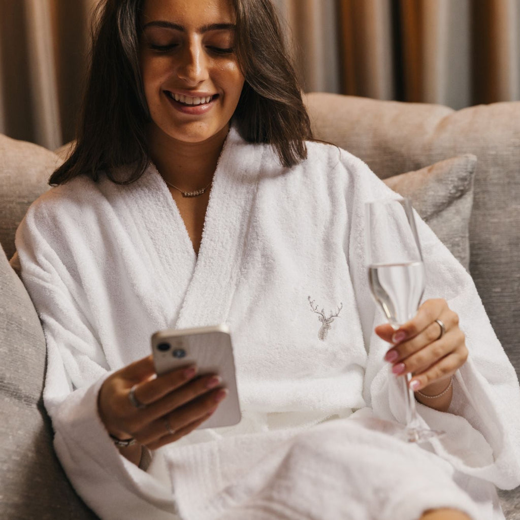 A tanned, brown haired woman is wearing a white pure 100% cotton terry towelling dressing gown from Loch Lomond Linen in an elegant hotel room and drinking chanmpagne while looking at her phone and smiling.